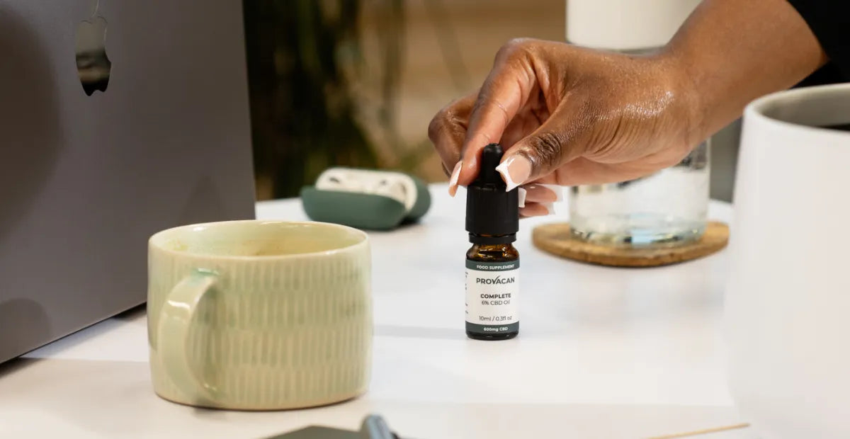 Person holding the dropper of a bottle of Provacan Complete 600mg 6% CBD oil on a desk with a mug, laptop and other items