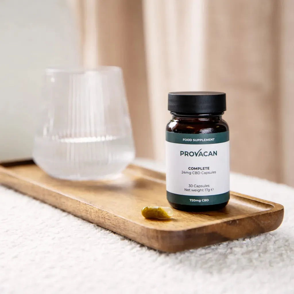 A jar of Provacan Complete 24mg CBD Capsules on a wooden tray next to a glass of water.
