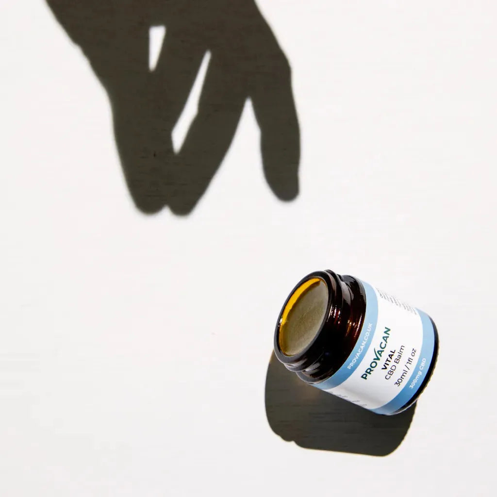 An open jar of Provacan Vital CBD Balm on white background with dark shadow and a shadow of a hand reaching for it.