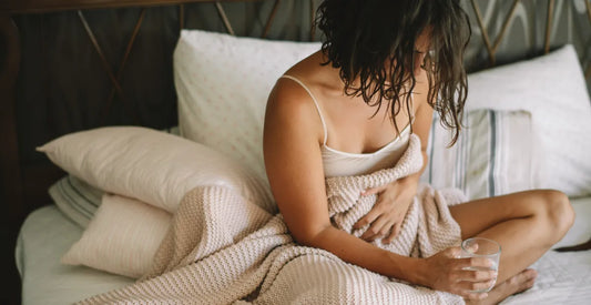 Woman on a bed visibly in pain, holding her belly with her hand and holding a glass of water in the other