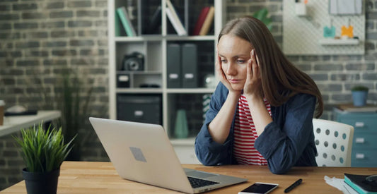 Woman in a denim jacket resting her head in her hands at her laptop, feeling the effects of stress on the body