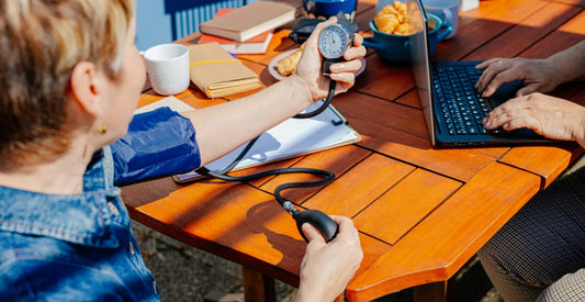Person measuring blood pressure while another person works on a laptop at a wooden table with coffee and notebooks
