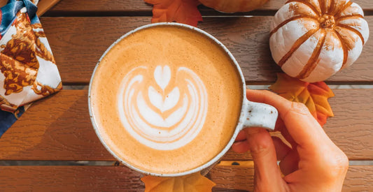 Person holding a cup of pumpkin spice latte with autumn-themed decor on a wooden table