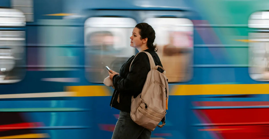 Woman in black jacket and jeans with a beige backpack holding her phone in front of a blue and yellow train passing by