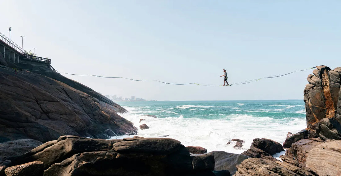 Person walking across a rope slackline between ocean cliffs under a clear blue sky