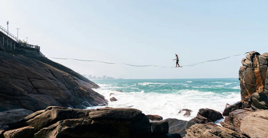 Person walking across a rope slackline between ocean cliffs under a clear blue sky