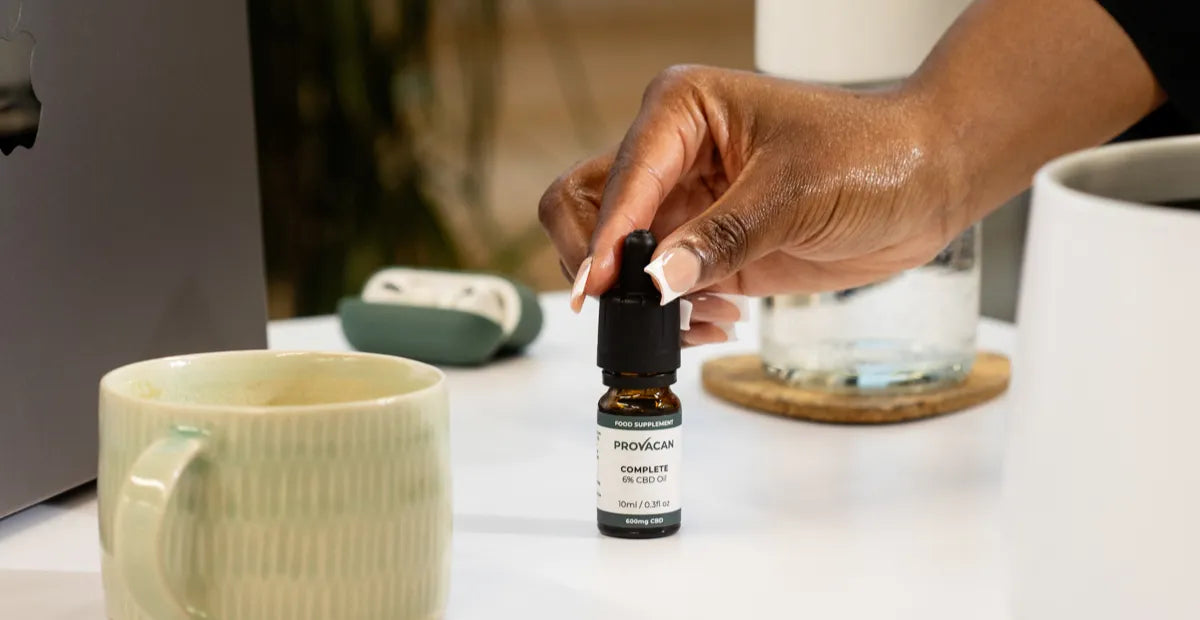 Woman's hand holding the dropper of a bottle of Provacan Complete 6% 600mg CBD oil on a desk