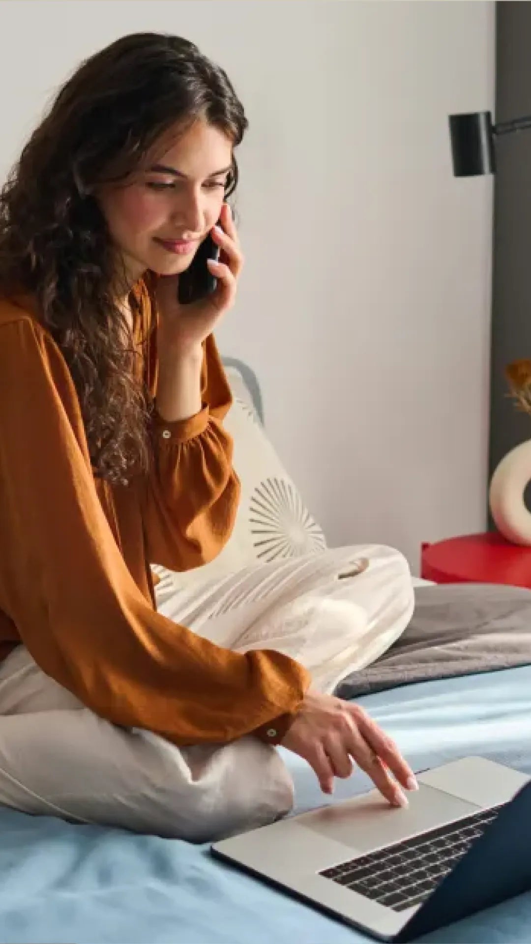 Woman sitting on a bed using a laptop with a phone to her ear