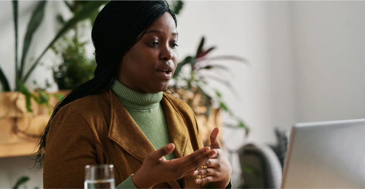 Woman in a brown jacket and green sweater sitting in a room with plants and a laptop