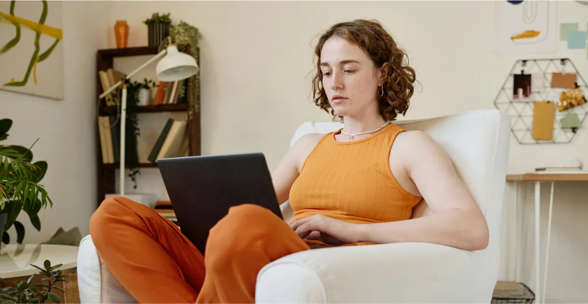 A young person with curly hair, wearing an orange tank top and orange pants, sitting in a white chair and looking at a laptop