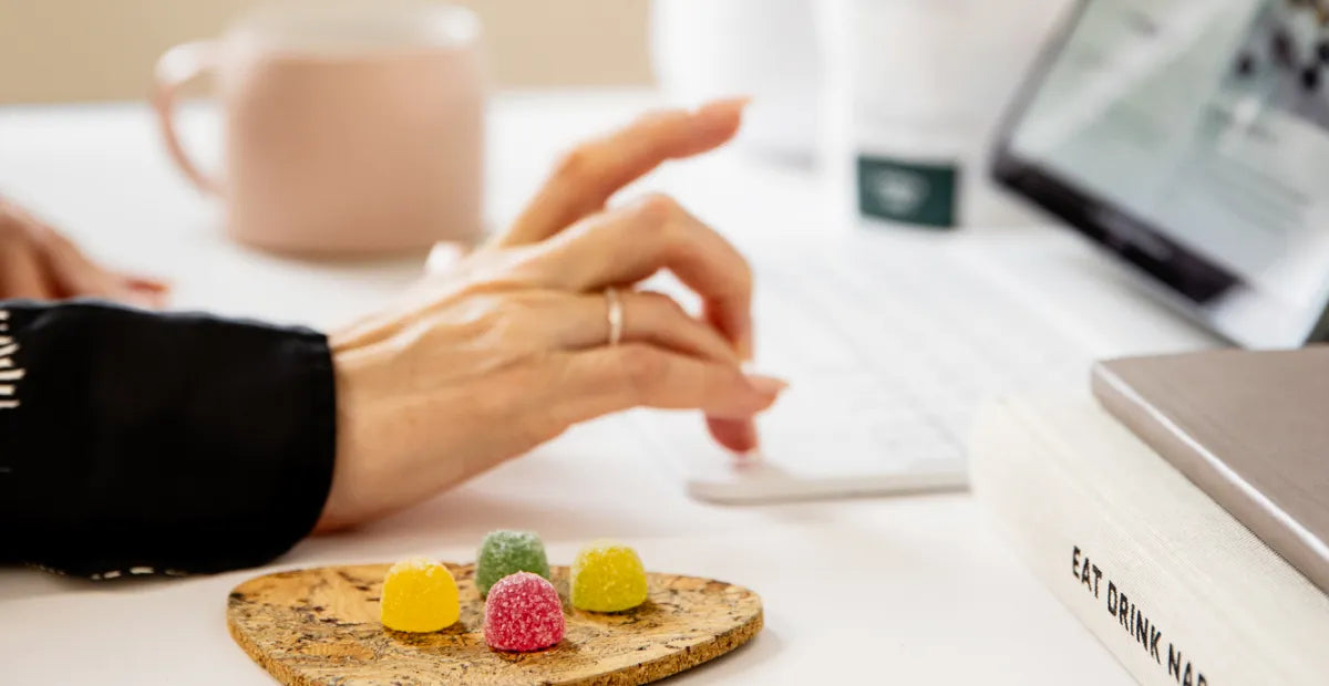 Person's hand typing on a laptop's keyboard on a desk with Provacan vegan CBD gummies on a tray at the front and mug in the background