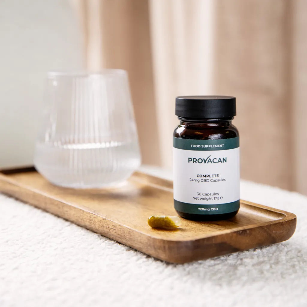 A jar of Provacan Complete 24mg CBD Capsules on a wooden tray next to a glass of water. 