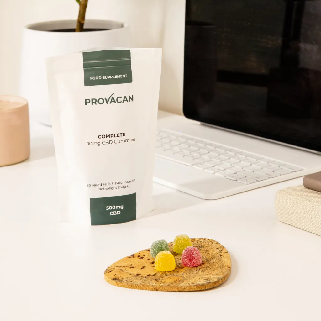 An open pack of Provacan Complete CBD Gummies on a desk, next to a laptop. A few colourful gummies are laid out on a tray next to the pack. 