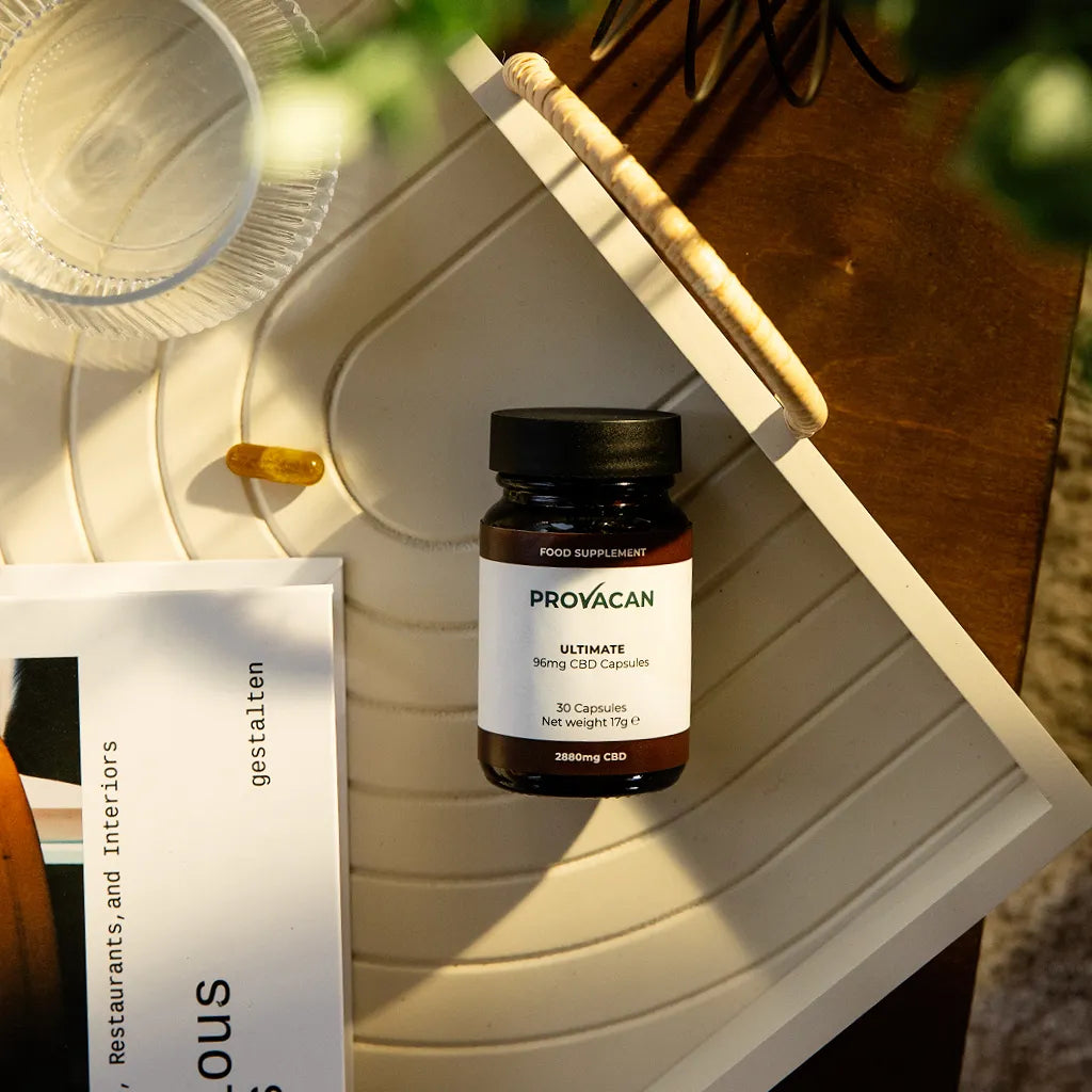 A jar of Provacan Ultimate 96mg CBD Capsules on a wooden tray with capsule, magazine, glass of water and plant in frame. 