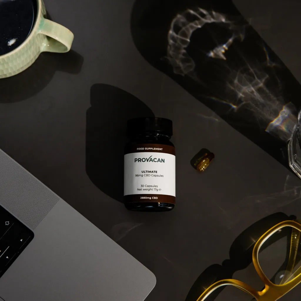 A jar of Provacan Ultimate 96mg CBD Capsules on a desk surrounded by coffee mug, laptop, glasses and glass of water. 